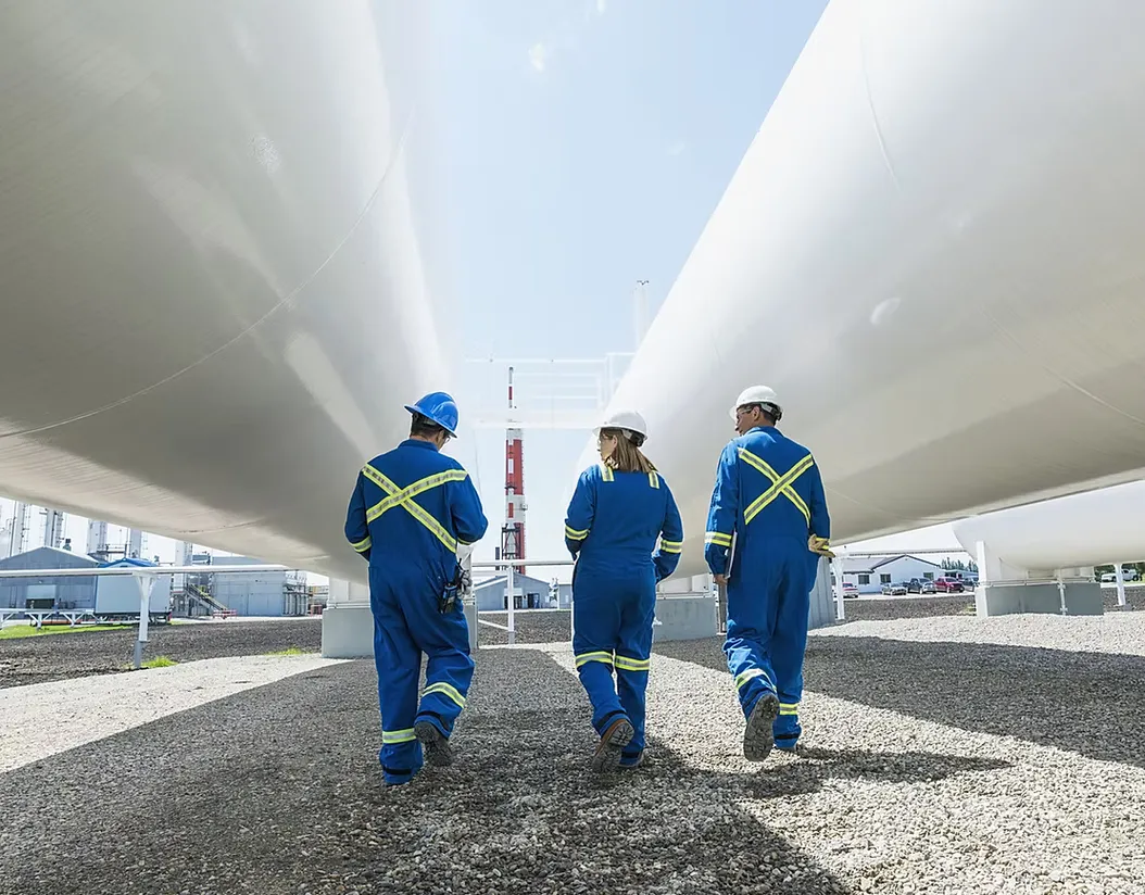 Three workers in blue coveralls with reflective tape and hard hats walk along a gravel path between large white horizontal storage tanks.