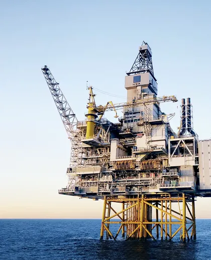 Offshore platform with a derrick, cranes and yellow steel jacket legs standing in dark blue sea under a clear sky.