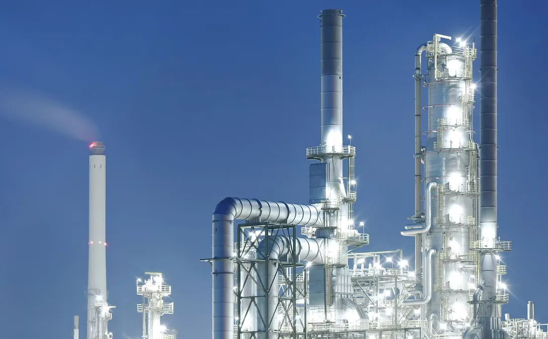 Illuminated refinery distillation columns and insulated pipework against a deep blue evening sky, with a smoking stack on the left.