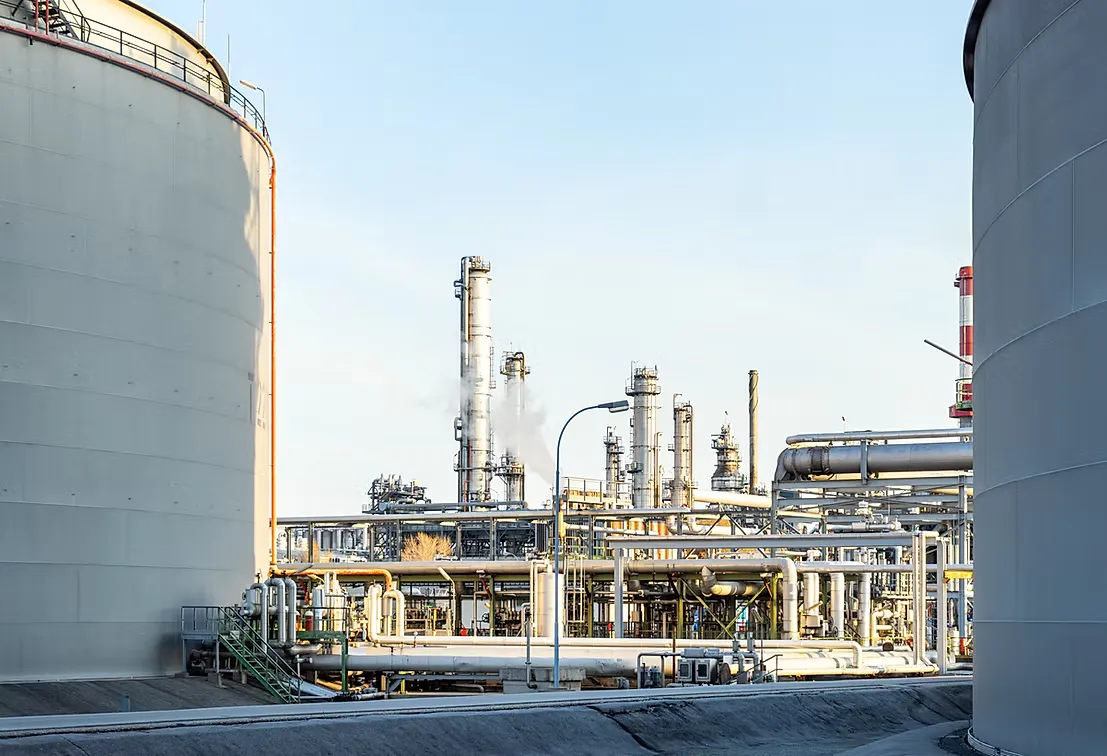 Refinery distillation columns and pipe racks seen between two large white storage tanks under a pale blue sky.