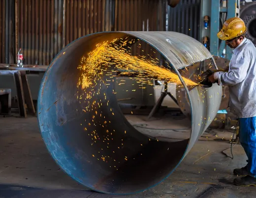 Worker in a yellow hard hat grinds the edge of a large rolled steel cylinder, sending orange sparks across its inside, in a dim workshop.